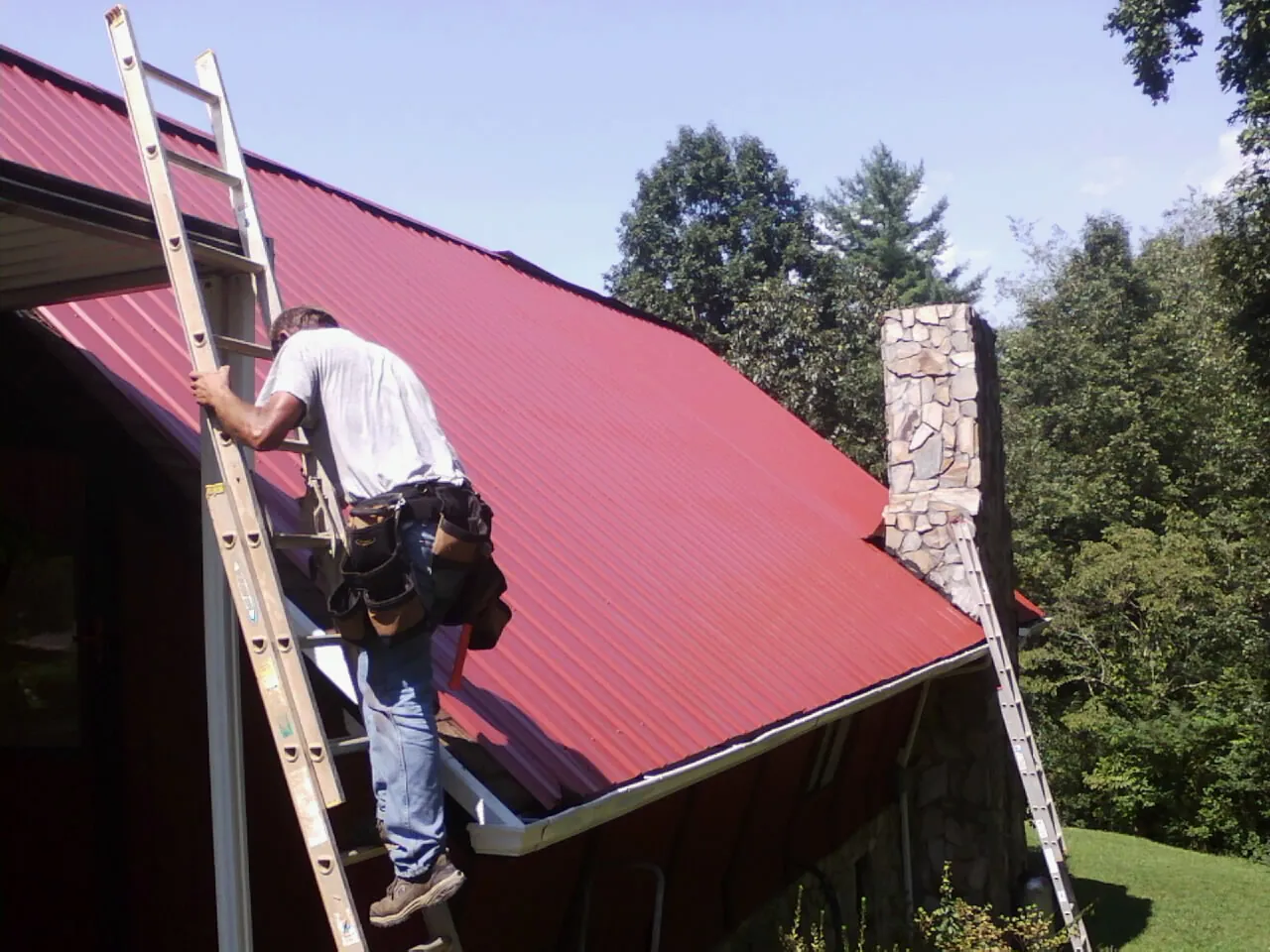 Roofer on a ladder beside a red metal roof and stone chimney — Soffit & Fascia Repair by Roofing Brothers in Wagener